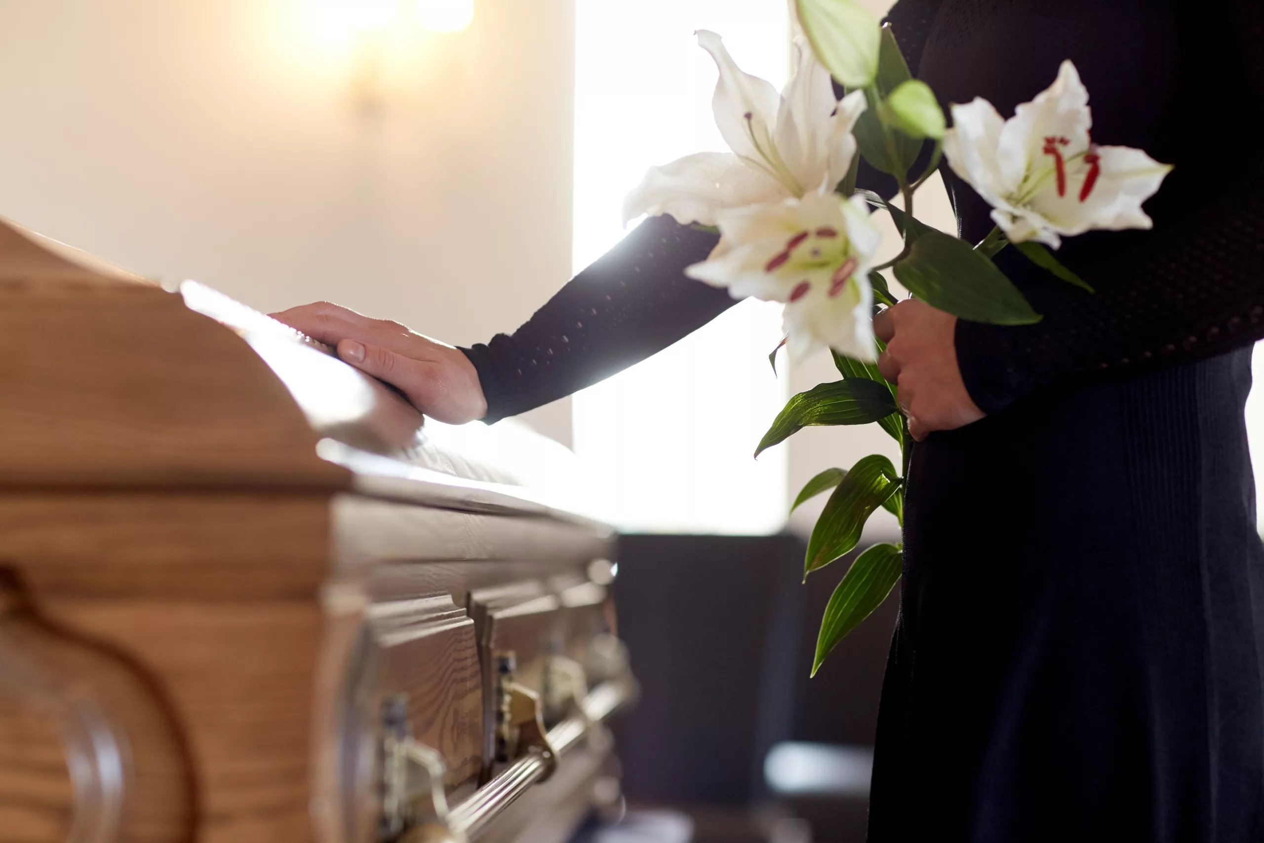 Hand on wooden casket holding white lilies, conveying mourning and respect in wrongful death claim context in Georgia